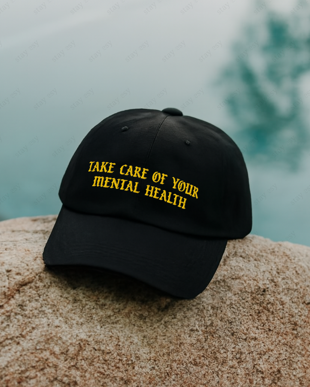 Black cap with 'Take care of your mental health' text on a rock by a body of water.
