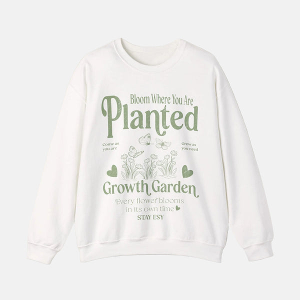 A white crewneck sweatshirt with a green graphic print on the front that reads 'Bloom Where You Are Planted Growth Garden' with small illustrations of flowers, hearts, and butterflies, shown against a light beige background.