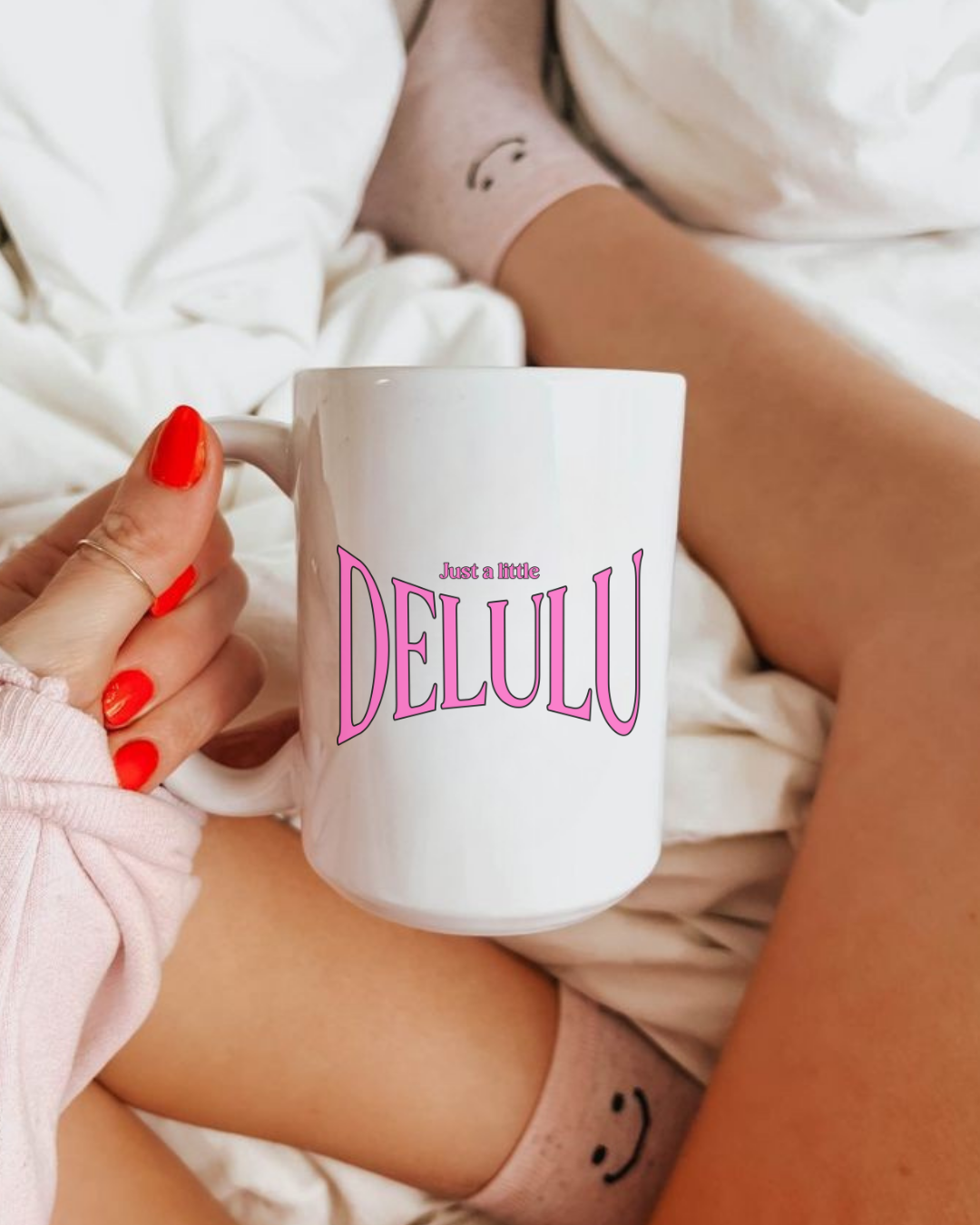 Person holding a white mug with 'DELULU' printed on it, sitting on a white surface.