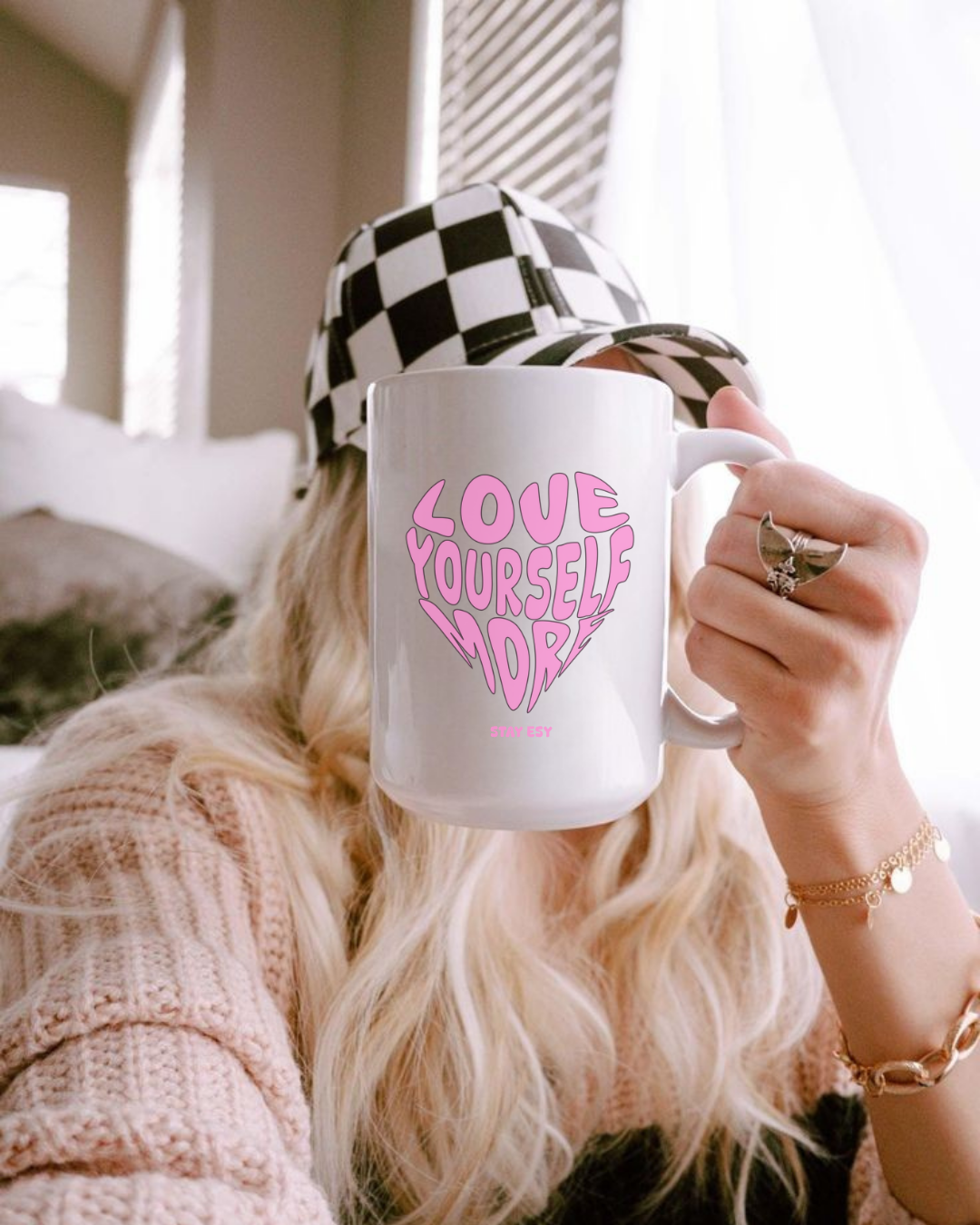 Person holding a mug with 'Love Yourself' text, wearing a checkered hat indoors.