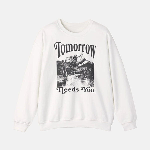 A white crewneck sweatshirt with a black graphic print on the front featuring the text 'Tomorrow Needs You' and a mountain landscape illustration, displayed against a light beige background.
