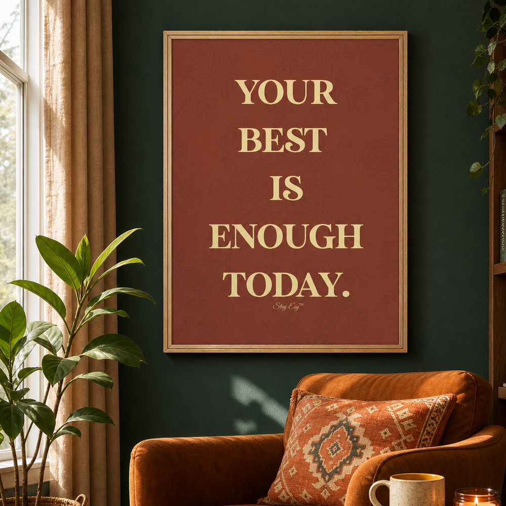 A framed motivational wall art print with the text 'YOUR BEST IS ENOUGH TODAY.' in cream lettering on a muted reddish-brown background, hanging on a dark green wall above a rust-orange armchair in a styled living room.