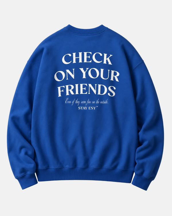 A royal blue crewneck sweatshirt shown from the back, featuring the white printed text 'CHECK ON YOUR FRIENDS' with smaller script text underneath, laid flat on a light beige background.