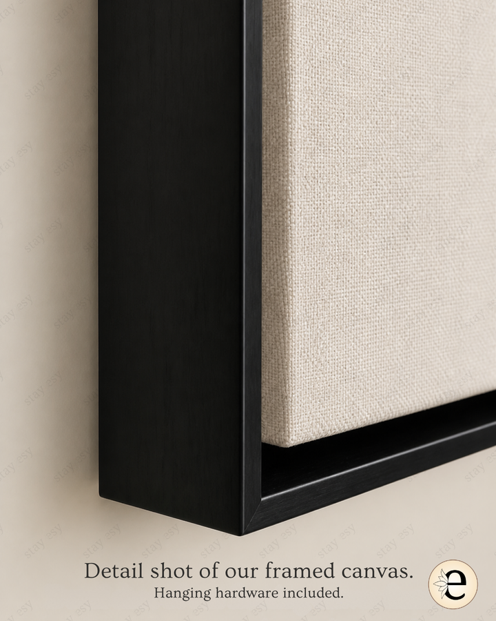 Close-up detail shot of a framed canvas wall art piece, showing a black frame and textured off-white fabric panel against a beige wall, with text reading &