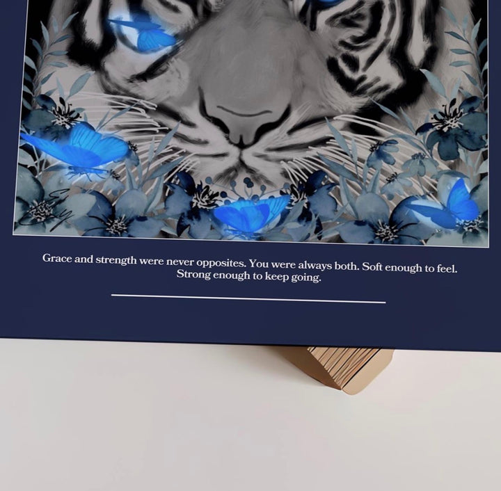 A square art print or card featuring a stylized black-and-white tiger face surrounded by blue flowers and butterflies, with a quote printed below on a dark navy background. The print is photographed resting on a small wooden stand against a light neutral surface.