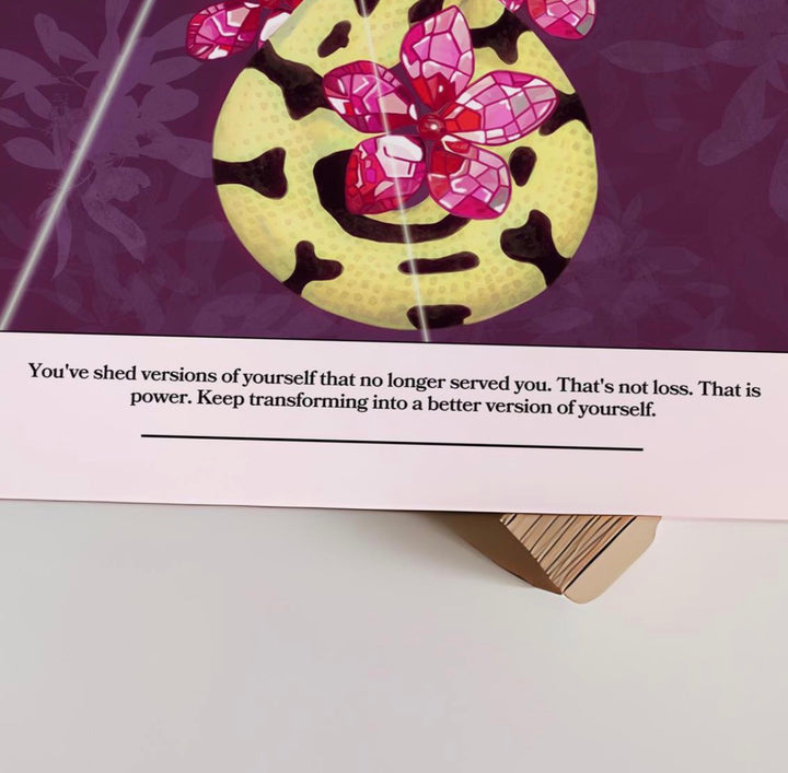 A close-up of a printed card or poster showing a purple background with a stylized yellow circular face, pink gem-like flowers, and a motivational quote on a white band below.