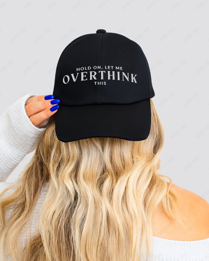 Black cap with 'Hold on, let me overthink this' text held by a person with blonde hair.
