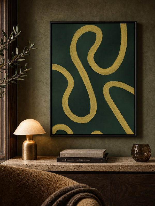 Framed abstract wall art featuring bold mustard yellow curved lines on a dark green background, displayed in a warm, rustic room setting above a console table with a lamp, books, and a textured vase.