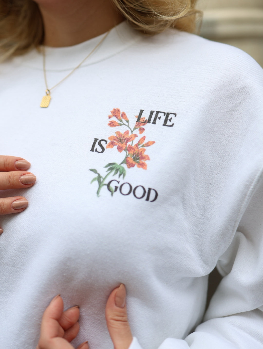 White sweatshirt with 'Life is Good' text and floral design worn by a person.