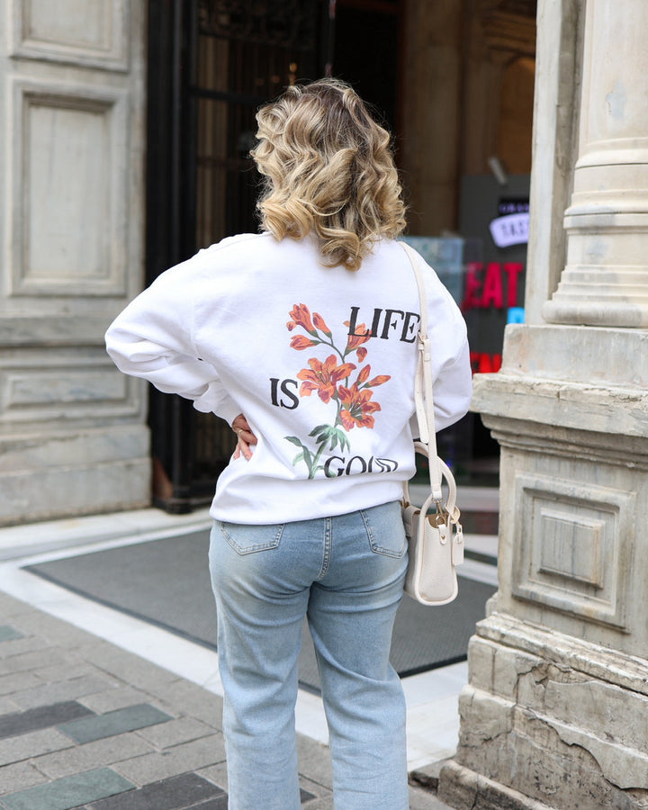 Person wearing a white sweatshirt with floral design and text, standing in front of a building entrance.