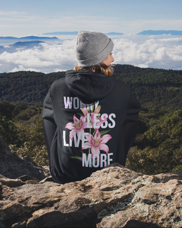 Person wearing a black hoodie with floral design and text, sitting on a mountain overlooking a scenic landscape.