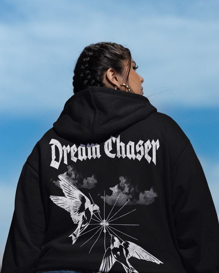 Person wearing a black hoodie with 'Dream Chaser' design against a blue sky.