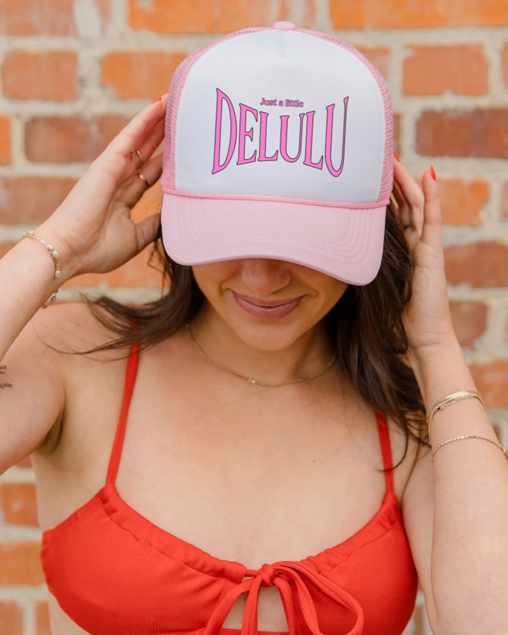 Person wearing a pink cap with 'Just a Little DELULU' text, standing against a brick wall.