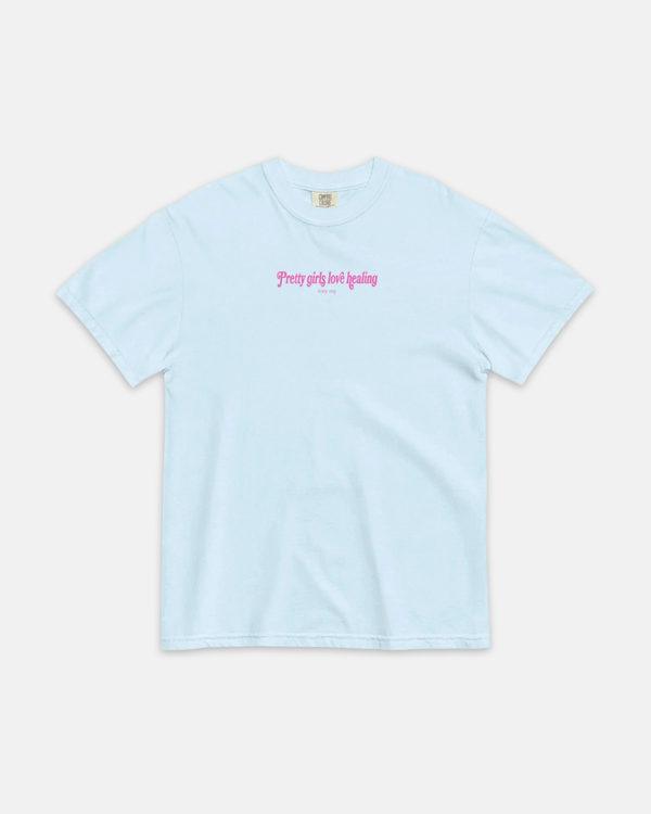 A light blue short-sleeve crewneck t-shirt with pink gothic-style text on the chest reading 'pretty girls just healing' and smaller text below, displayed flat against a light gray background.