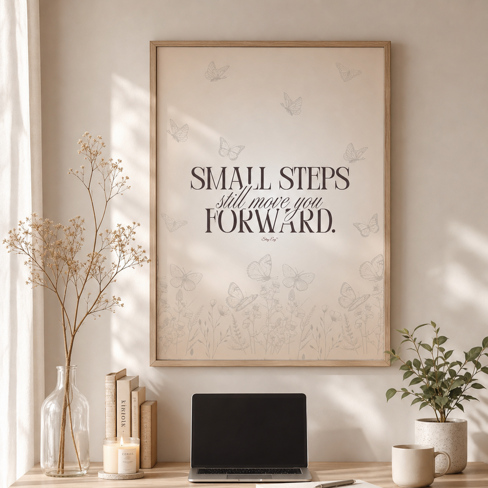 A framed motivational wall art print with the quote 'SMALL STEPS still move you FORWARD.' hangs above a wooden desk in a styled home office setting with a laptop, books, candles, and plants.