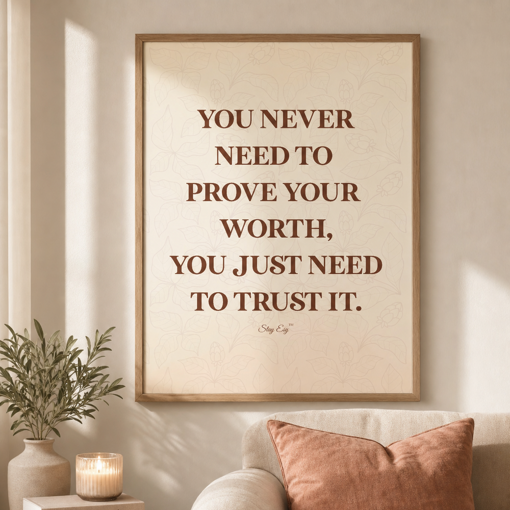 A framed motivational wall art print with the quote 'YOU NEVER NEED TO PROVE YOUR WORTH, YOU JUST NEED TO TRUST IT.' hangs above a beige sofa with a rust-colored pillow and a knit throw blanket in a softly lit living room.