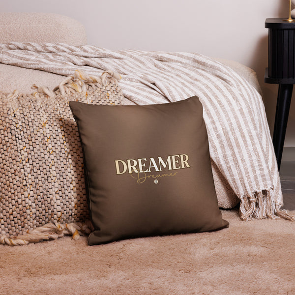 Brown pillow with 'DREAMER' text on a bed with beige bedding and blanket.
