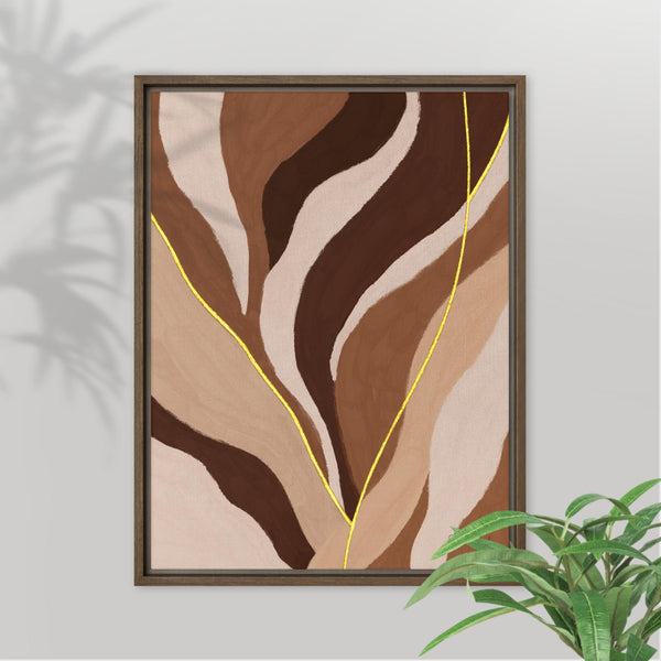 A framed abstract wall art print with flowing organic shapes in brown, tan, cream, and dark brown, accented by thin yellow lines, shown hanging on a light gray wall with a plant in the foreground.