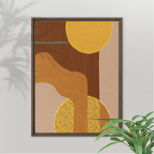 A framed abstract wall art print with warm earthy tones, featuring a large mustard yellow circle, brown and tan organic shapes, and a beige background, shown hanging on a light gray wall with a plant leaf in the foreground.