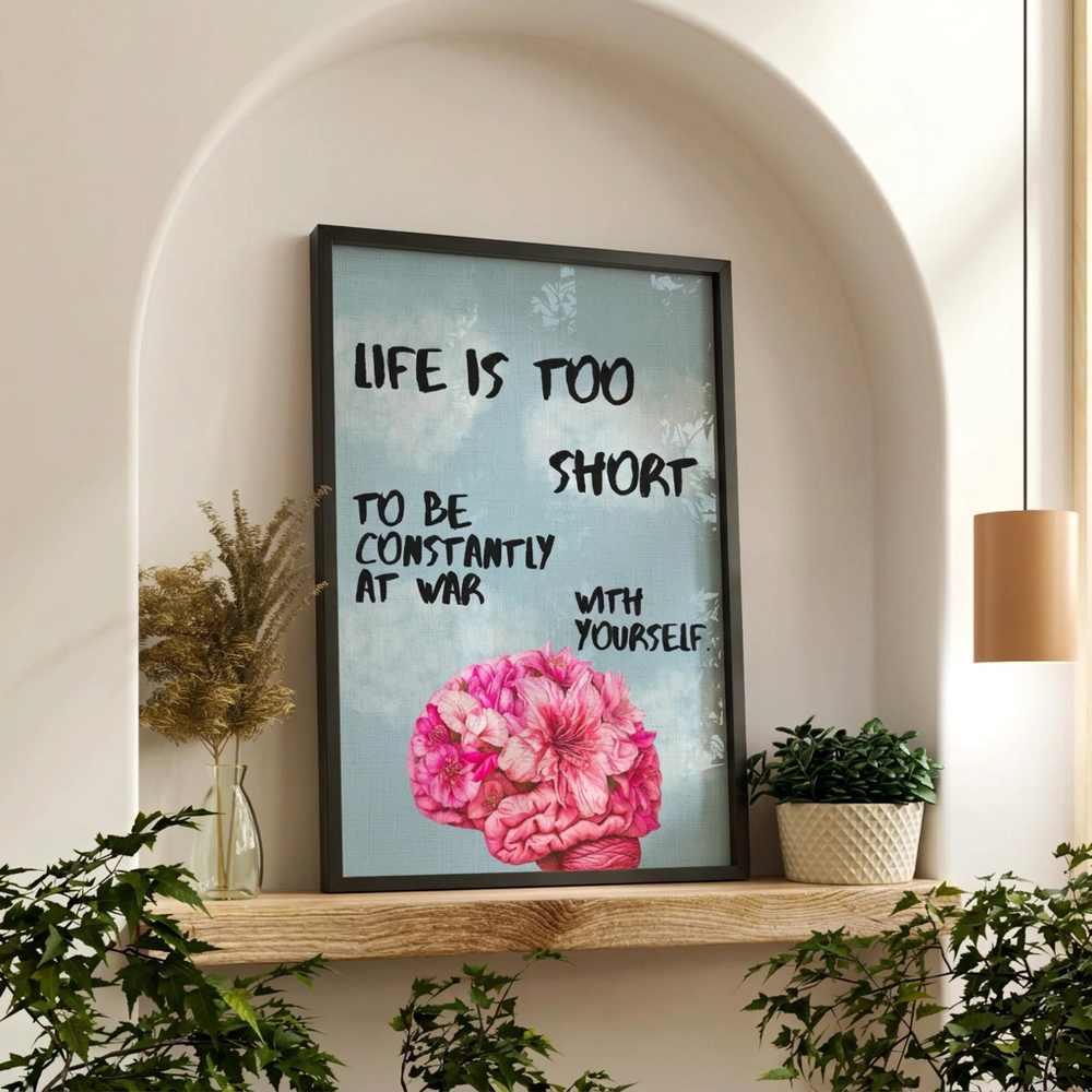 A framed wall art print with a motivational quote and a pink flower illustration, displayed on a wooden shelf in a styled interior setting with plants and a hanging lamp.