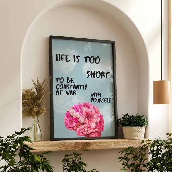 A framed wall art print with a motivational quote and a pink flower illustration, displayed on a wooden shelf in a styled interior setting with plants and a hanging lamp.