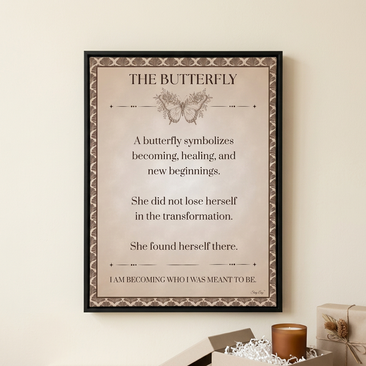 A framed typographic art print titled "THE BUTTERFLY" hangs on a beige wall above a styled surface with a candle and gift boxes. The print has a cream background, decorative border, a butterfly illustration, and centered text about transformation and self-discovery.