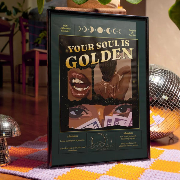 A framed art print with the text 'YOUR SOUL IS GOLDEN' and moon phase illustrations, styled on a patterned rug with disco ball props in a warm indoor setting.