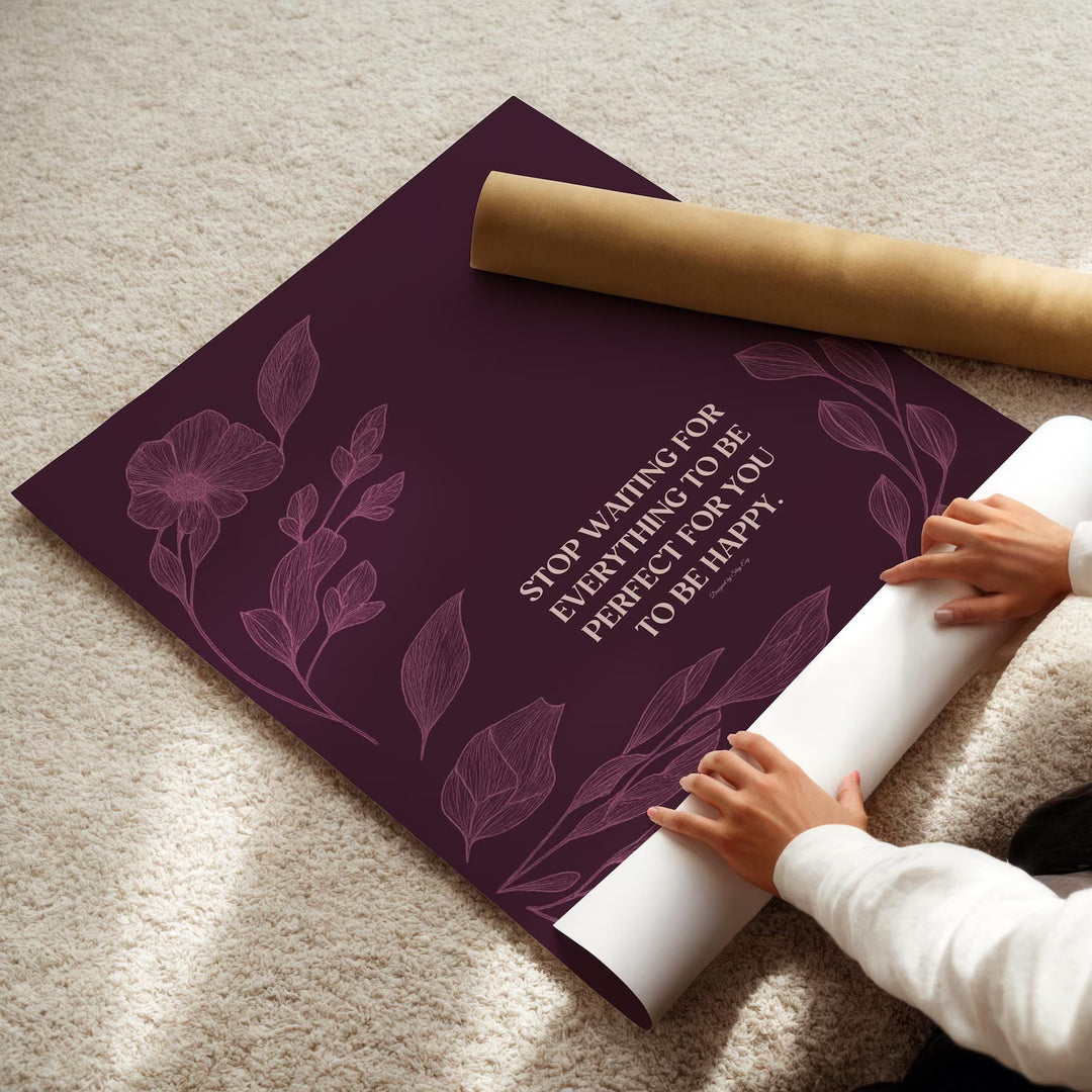 A person unrolls a large decorative poster or art print on a beige carpet. The print has a deep plum-purple background with floral line art and the text &
