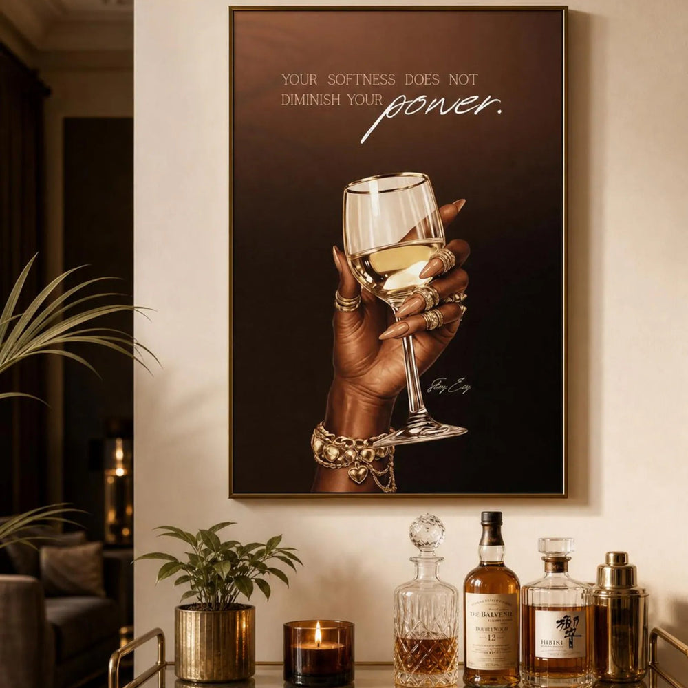 A framed wall art print hangs above a bar cart in a warm, stylish interior. The print shows a hand holding a wine glass against a dark brown background with the text, 'YOUR SOFTNESS DOES NOT DIMINISH YOUR power.