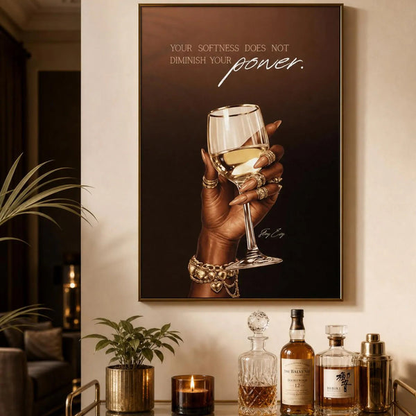 A framed wall art print hangs above a bar cart in a warm, stylish interior. The print shows a hand holding a wine glass against a dark brown background with the text, 'YOUR SOFTNESS DOES NOT DIMINISH YOUR power.
