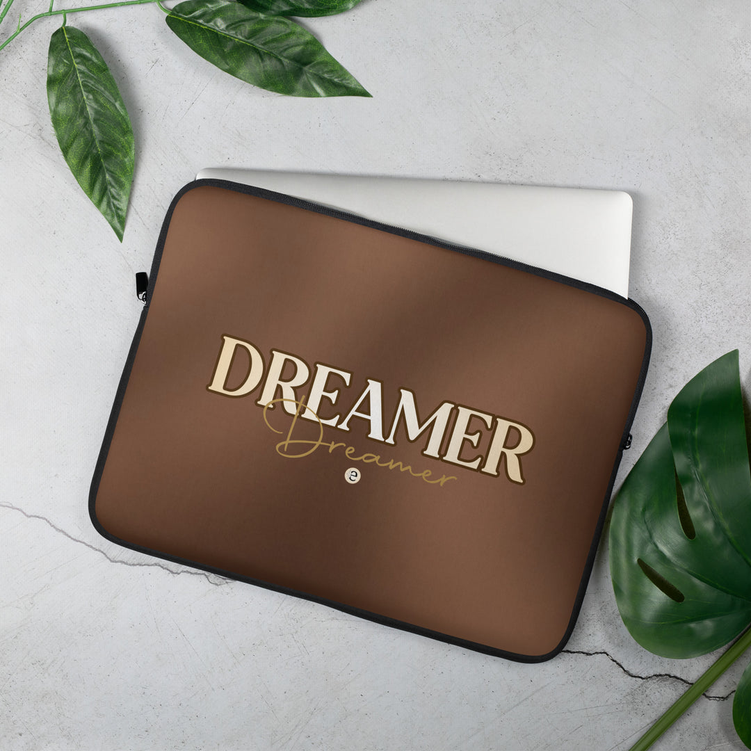 Brown laptop sleeve with 'DREAMER' text on a light gray surface with green leaves.