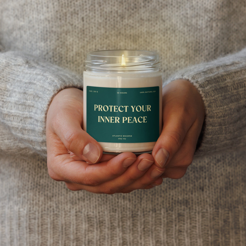 Candle with 'Protect Your Inner Peace' label held in hands against a textured fabric background