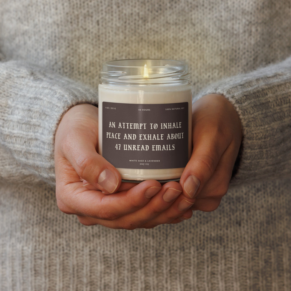 Candle in a jar held by hands with a humorous label, against a neutral background.