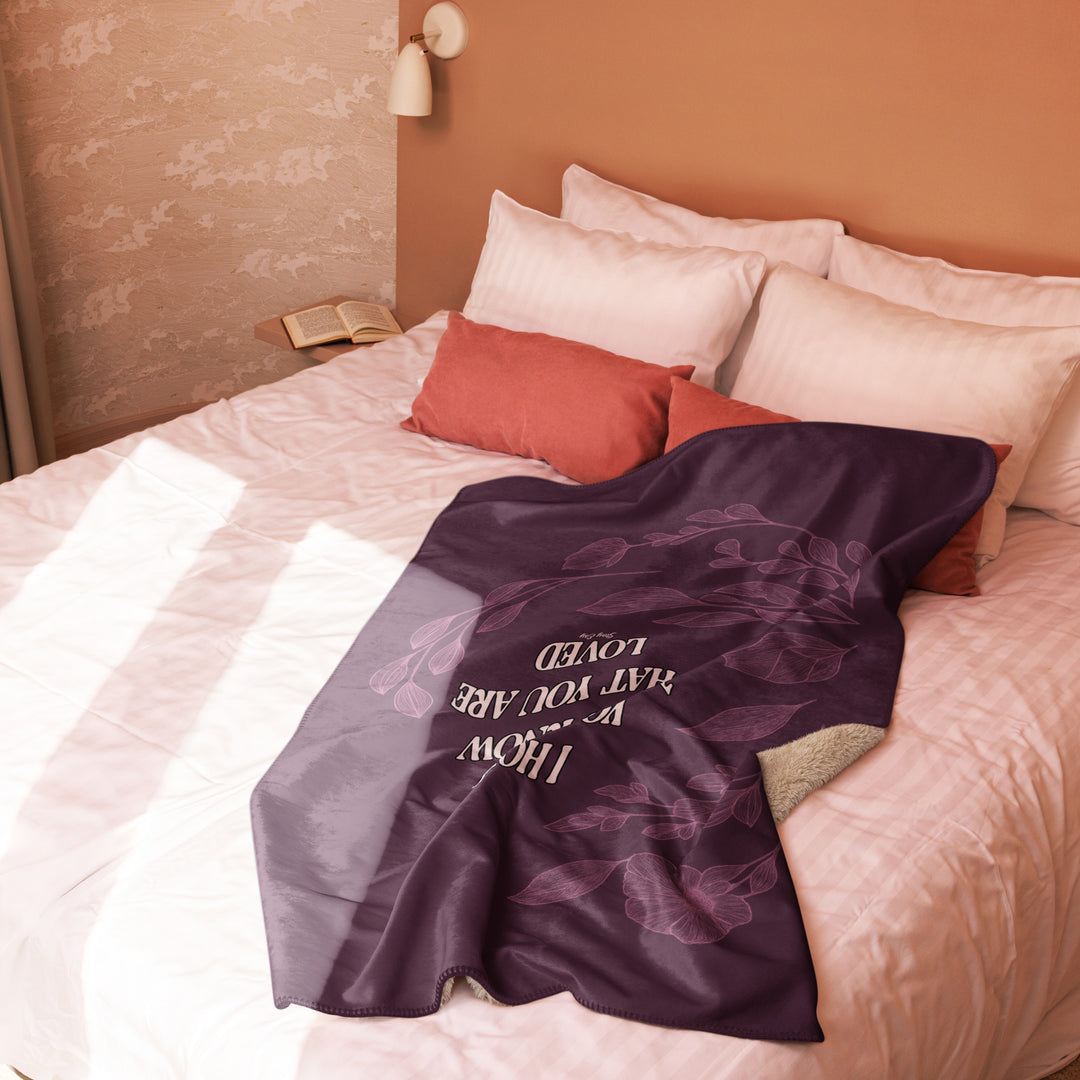 Purple blanket with text draped over a bed with pillows and a lamp in the background.